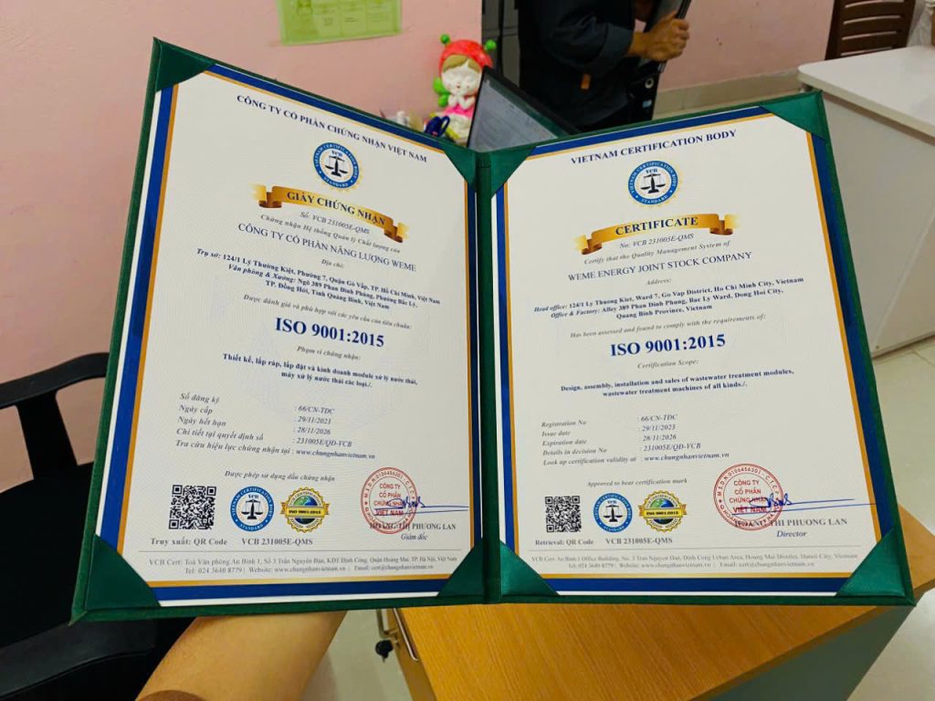 Chứng nhận ISO 9001:2015 song ngữ Việt – Anh của Công ty Cổ phần Năng lượng WeMe được đặt trong bìa hồ sơ, thể hiện tiêu chuẩn quản lý chất lượng cho hoạt động thiết kế, lắp ráp, lắp đặt và kinh doanh module xử lý nước thải