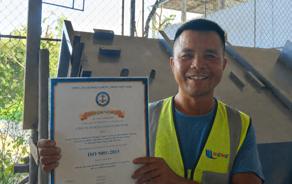 Nhân sự WeMe mặc áo bảo hộ lao động, cầm chứng nhận ISO 9001:2015 tại khu vực sản xuất module xử lý nước thải